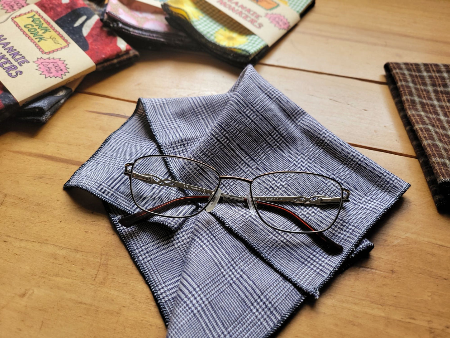 Eyeglasses on a plaid hankie with packaged hankies in the background on a wooden surface
