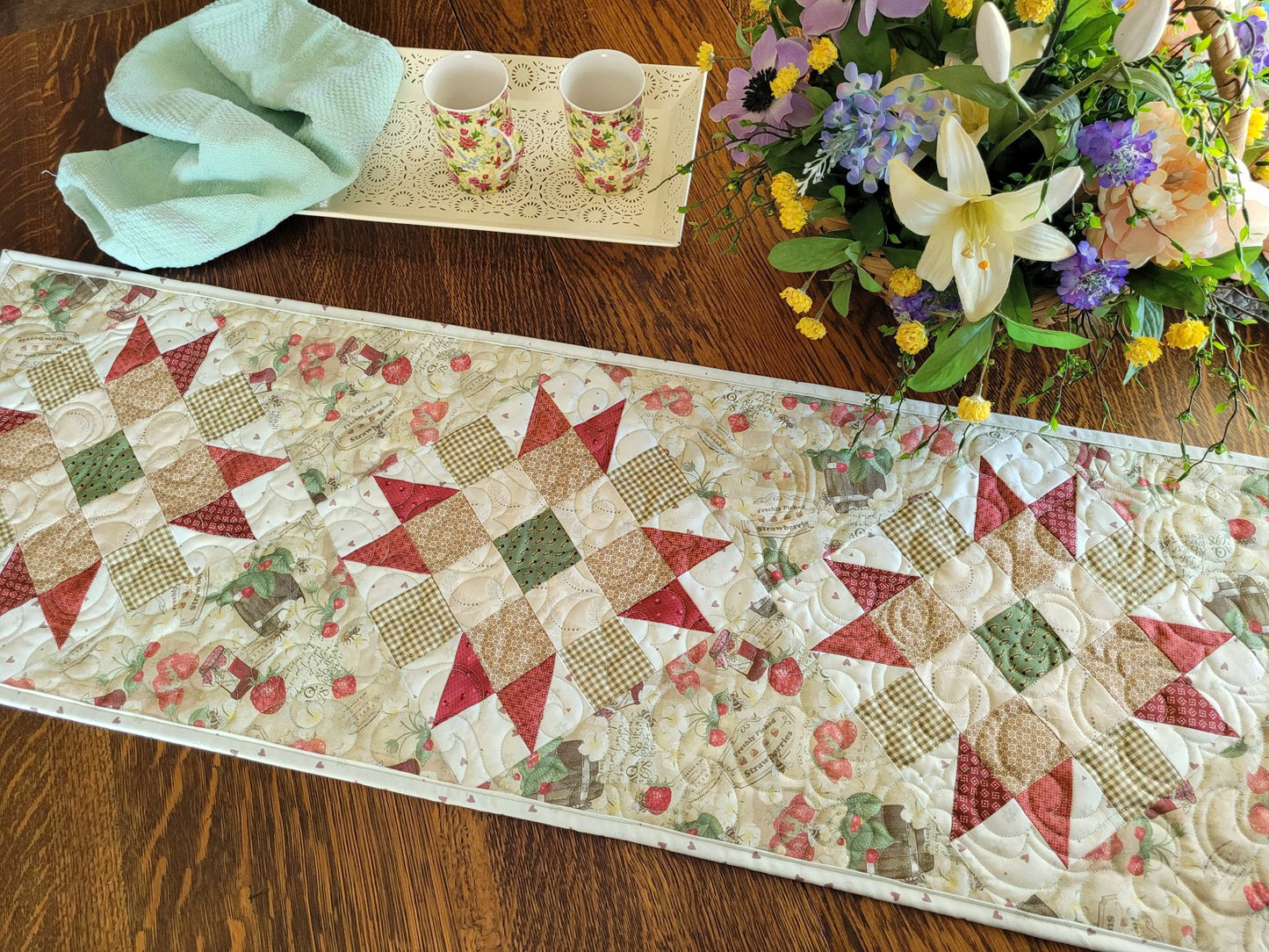 strawberry quilted table runner shown on table with a spring flower arrangement and tea tray with two mugs