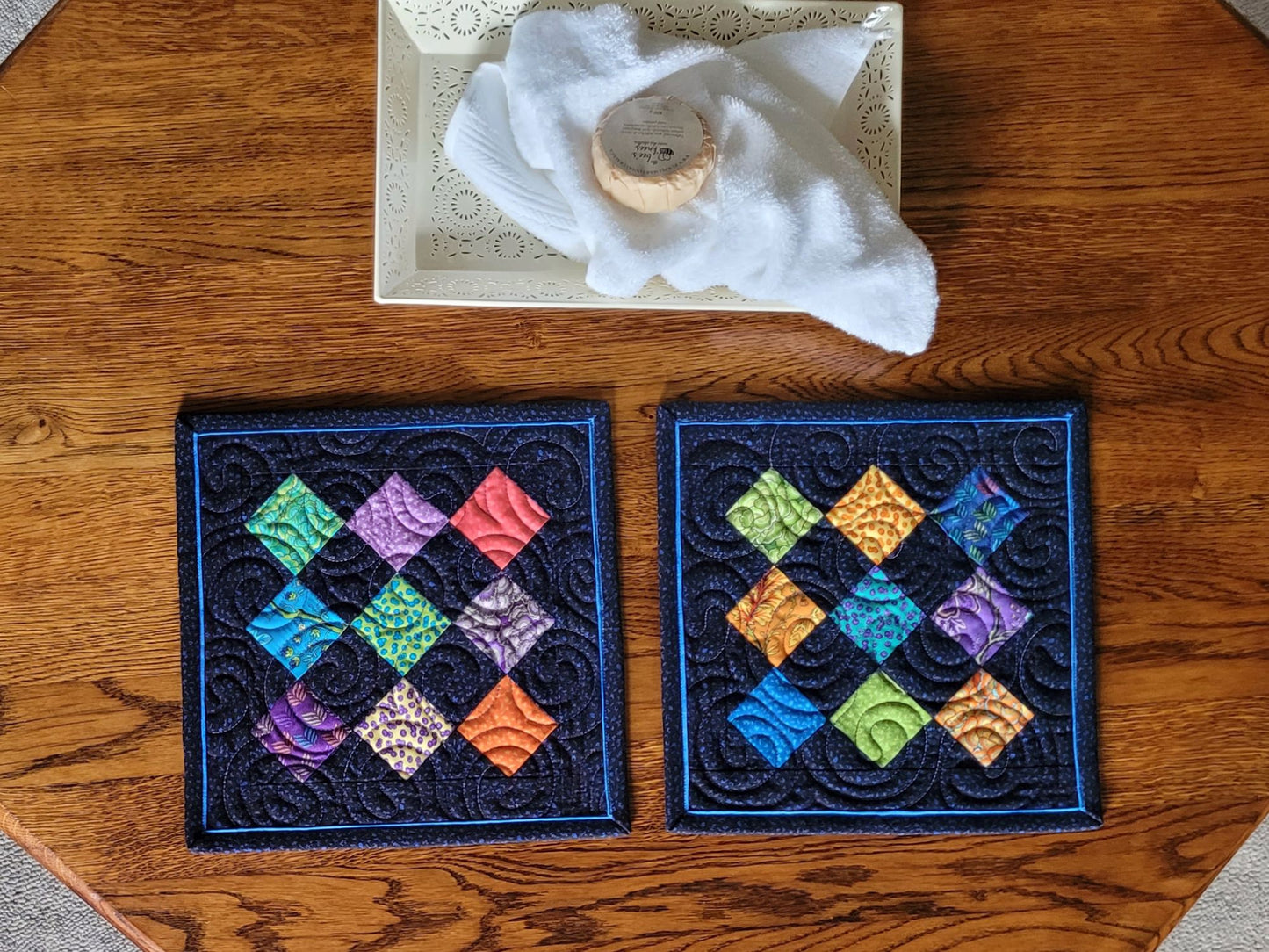 Two mini quilts with colourful diamond pattern on a wooden surface.