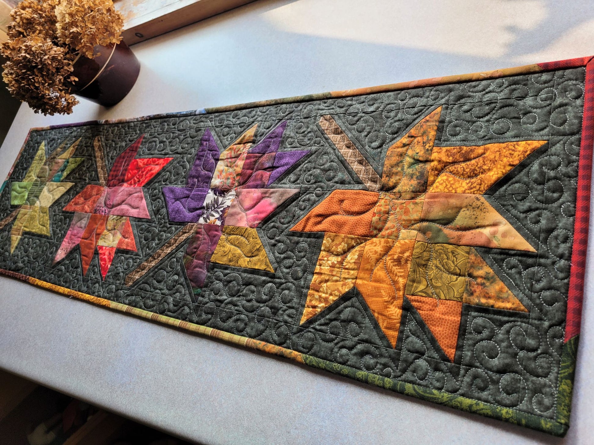 Quilted table runner with leaf patterns in autumn colours on a white surface.