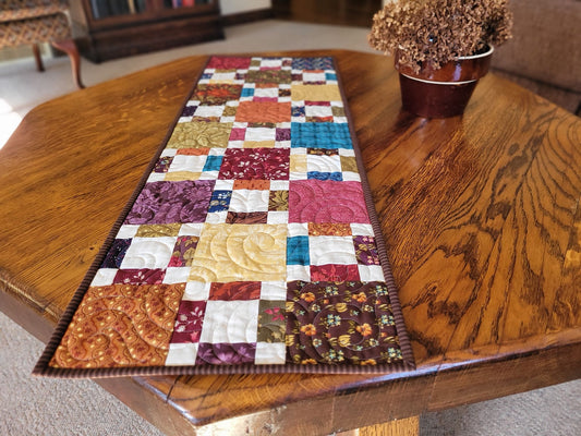 Colorful patchwork table runner on a wooden table with a vase of flowers in the background.
