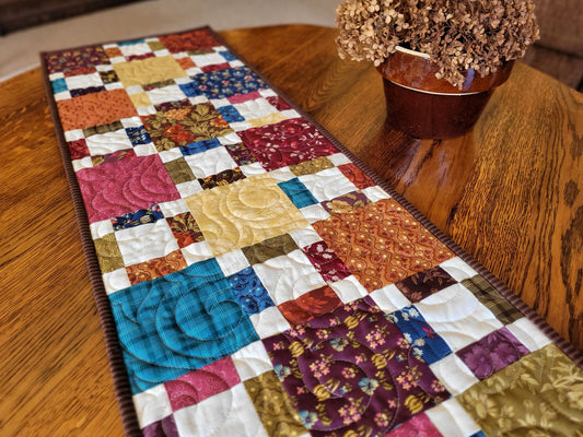 Colorful quilted table runner on a wooden table with a vase of flowers.