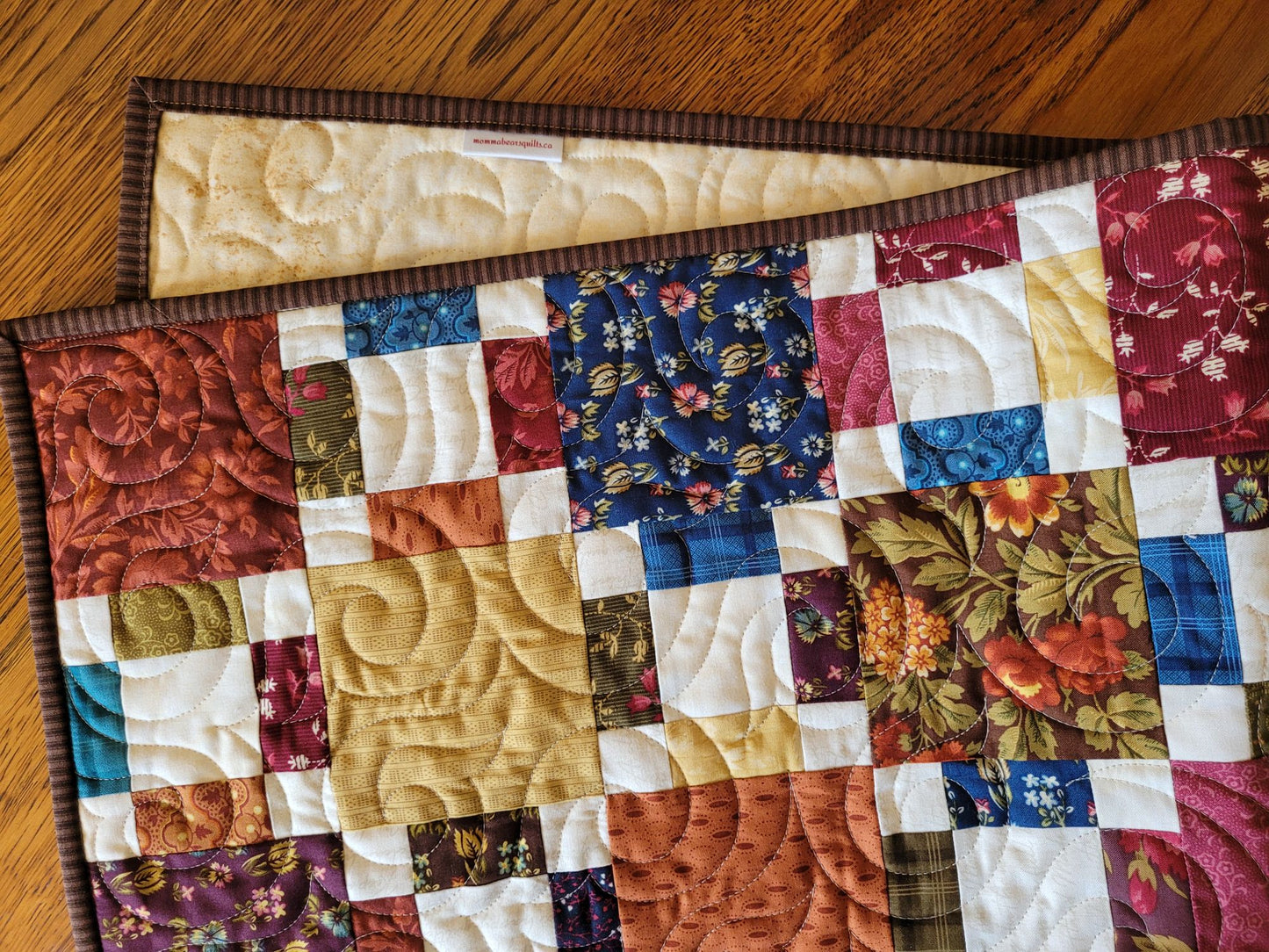 Quilted table runner with patchwork design on a wooden surface, showing a peek of backing.