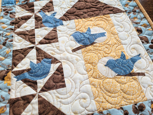 Close up of quilted wall hanging with blue birds flitting around a birdhouse. Warm white background shows freemotion quilting in a floral design.