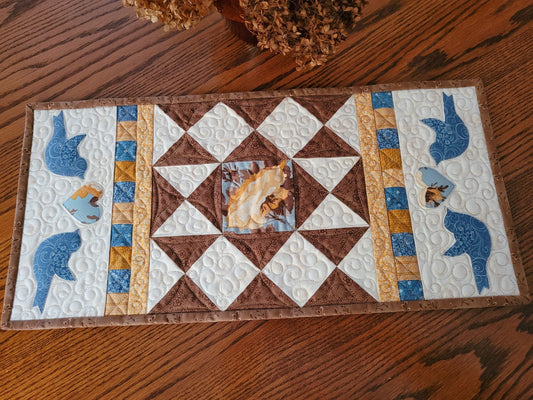 Quilted patchwork table runner on a wood table with dried flowers. On each end the runner has two sets of blue birds, each flanking a floral heart. The warm white background shows off the other coordinating fabric colours of brown, blue, gold and yellow.