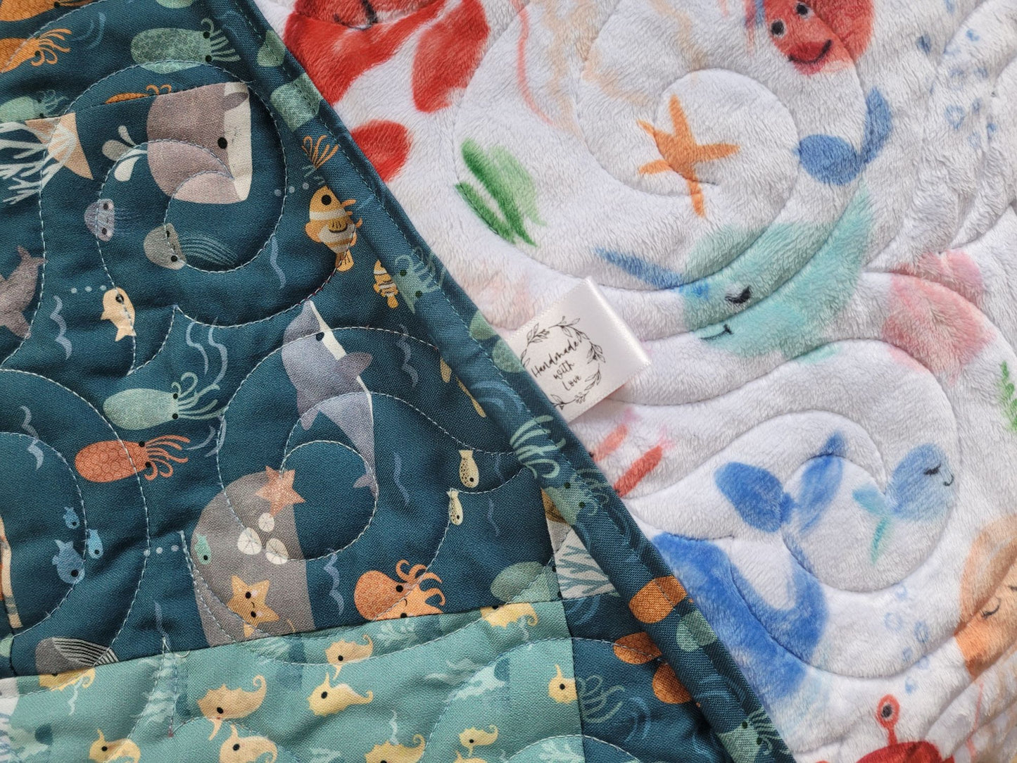 Close-up of baby quilt with sea-themed prints in dark and light teal with ocean animals like whales, dolphins, fish, and seahorses. Quilt is folded to show soft minky back with colourful crabs, starfish, and sea creatures. Handmade with Love label is shown in the centre of the photo.
