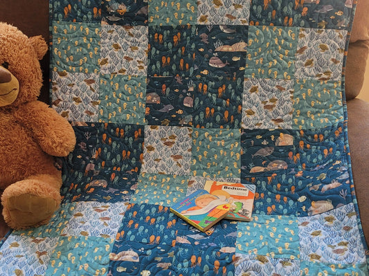 A handmade patchwork quilt featuring ocean-themed prints of whales, fish, and sea plants in shades of blue, teal, and white, styled with a brown teddy bear and a children's book on a couch.