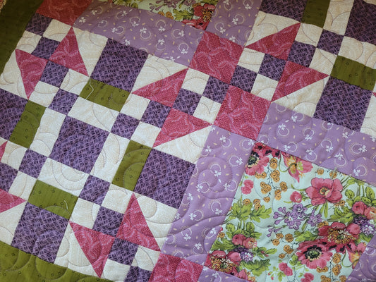 A close-up of a handmade quilt featuring a patchwork design with purple floral and patterned squares, pink and white checkered triangles, moss green blocks, and a floral print patch.