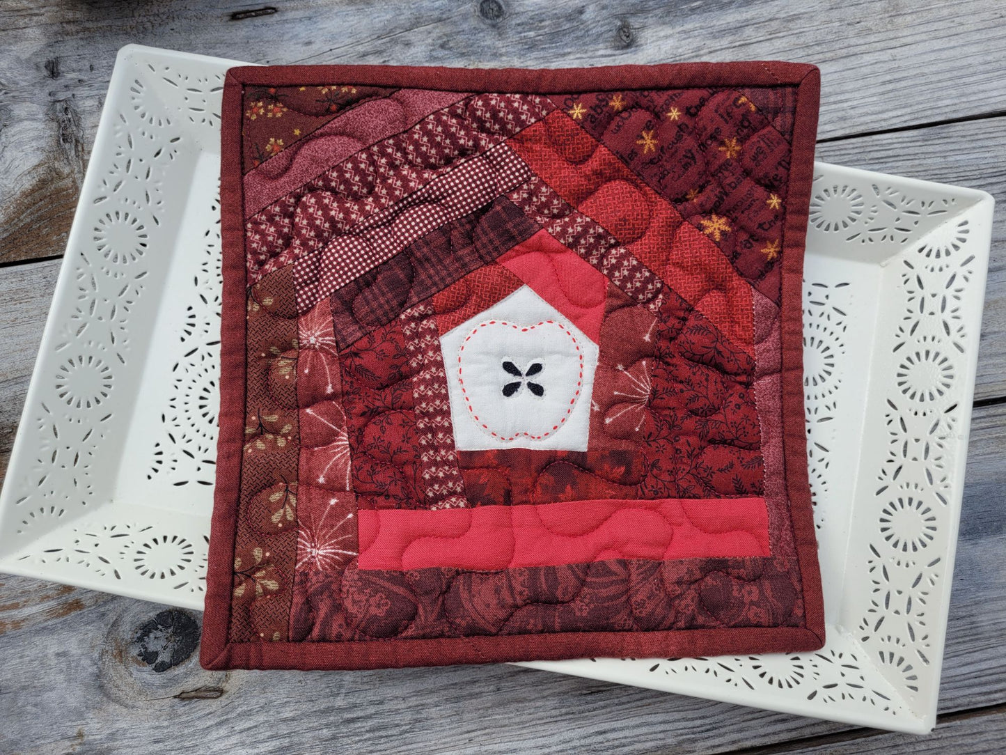 A square quilted patchwork hot mat featuring a handstitched apple design in the centre. Scrappy patchwork is in shades of red, burgundy, and white, displayed on a white ceramic tray over a rustic gray wooden surface.