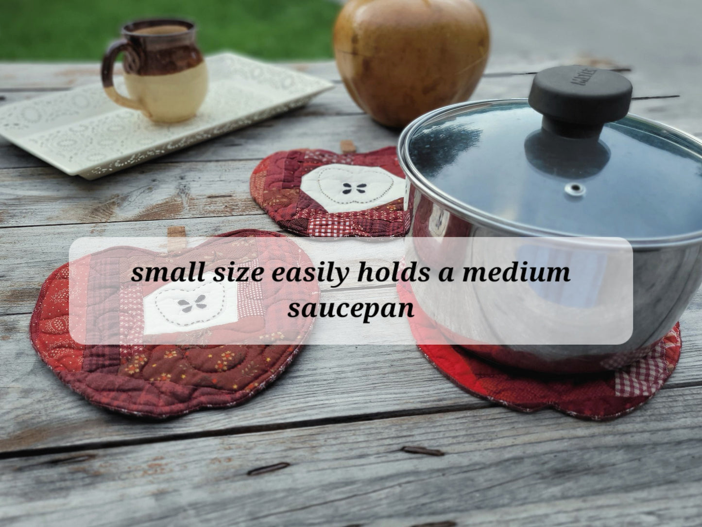 A stainless steel saucepan with a glass lid sits on a rustic wooden table, resting on a red patterned quilte apple hot mat. A text overlay reads 'small size easily holds a medium saucepan'.