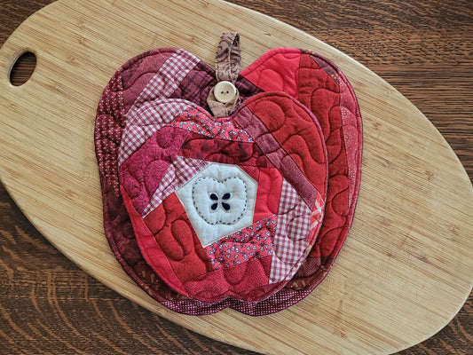 An apple-shaped quilted potholder with an apple design, featuring red and burgundy fabrics, resting on a light-colored wooden cutting board over a dark wood table.