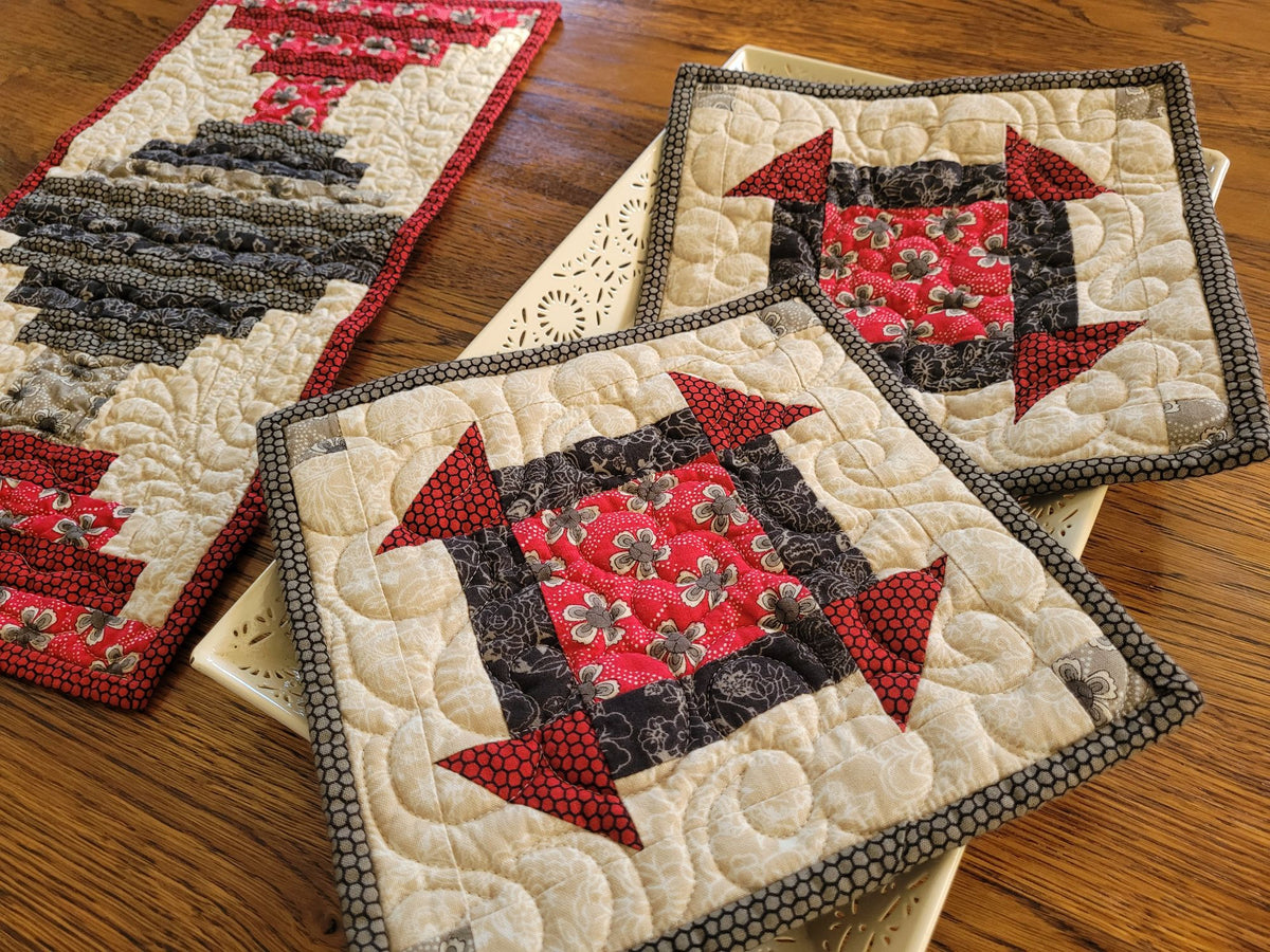 Three quilts arranged on a wooden table. The two hot mats are laying on a white decorative tray with the small table runner in the background on the left side. The coordinating pieces feature a patchwork design with red, black, gray and cream fabrics in floral and geometric patterns.