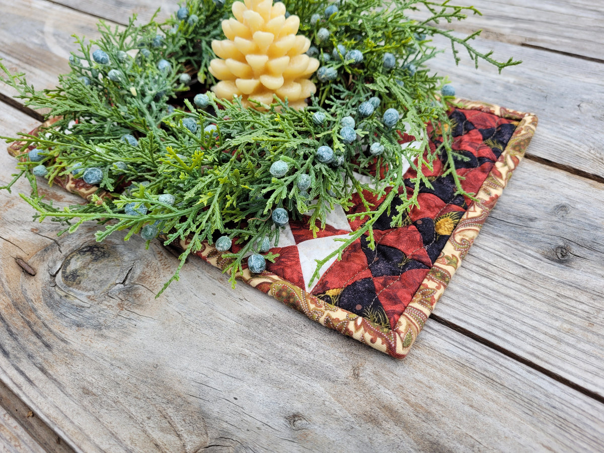 Christmas greenery with a candle in the middle is shown in a small patchwork quilt on a weathered wooden table.