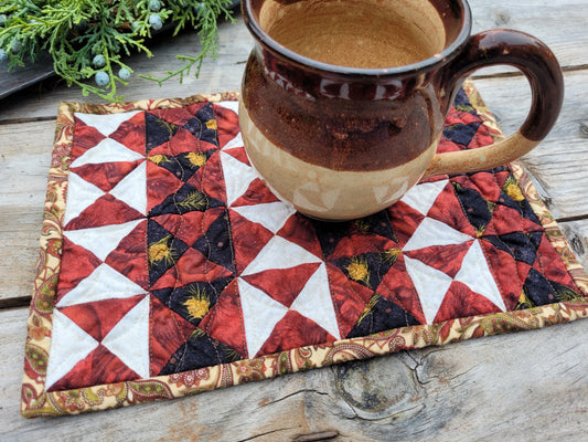 Close up of a small red and black quilted mat shown on a weathered wooden table with a brown pottery mug sitting on it. Christmas greenery is partially visable in the upper left corner.