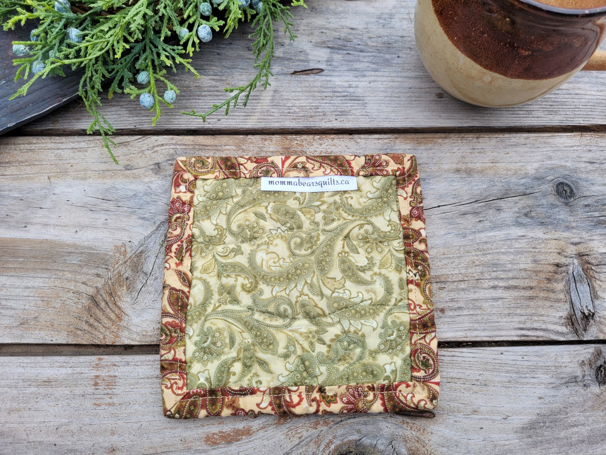 Showing the back of a quilted coaster on a rustic wooden table with a mug and greenery partially visable.