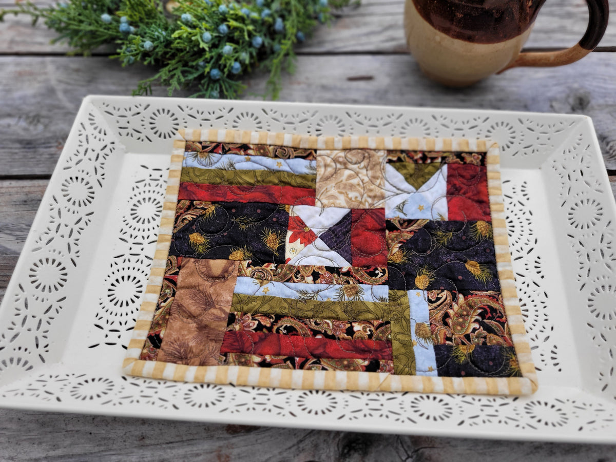 A small quilted fabric mat with a patchwork design in red, brown, black, beige, and light blue, featuring a striped yellow and white border. The quilt is displayed on a white decorative tray on a rustic wooden table, with a mug and greenery in the background.