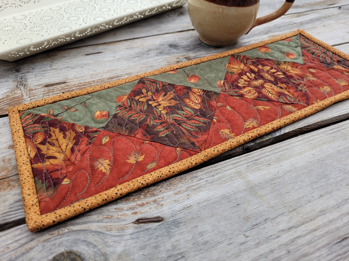 A small skinny quilt in rustic fall leaves and rich colours of burnt orange, deep red and sage green is shown on a weathered wooden table with a pottery mug and white decorative tray partially visable on the top left.