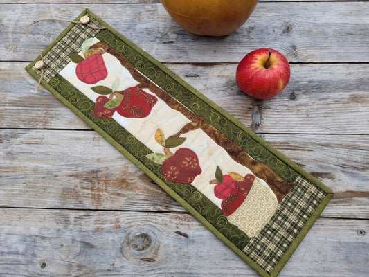 A skinny wall quilt is laying diagonally on a weathered wood table with a red apple nearby and a gourd partially visable. The quilt features applique apples with green patterened borders.
