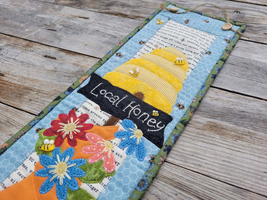 A long narrow quilt featuring a bee skep, a sign saying 'local honey' and daisy-like flowers is shown partially visable on a weathered wood table.