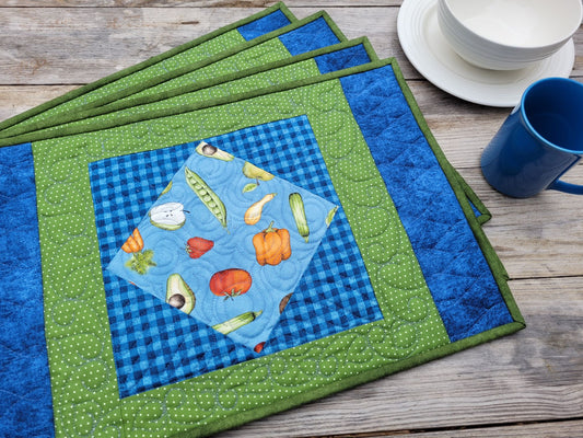 A set of handmade quilted fabric placemats arranged on a rustic wooden table, featuring green and blue borders with a central blue gingham and vegetable print square.