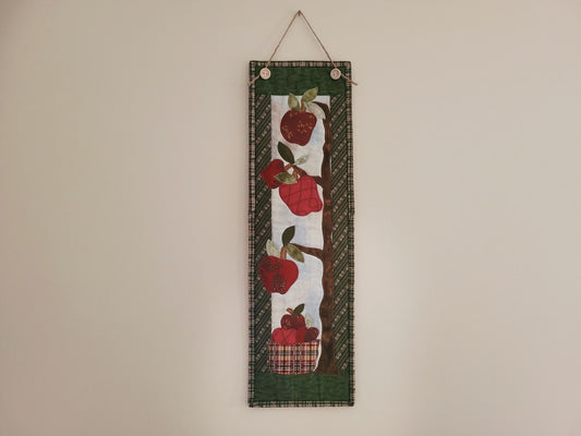 A skinny wall quilt featuring apples hanging from tree branches with a basket of apples underneath is shown hanging on a plain beige wall.