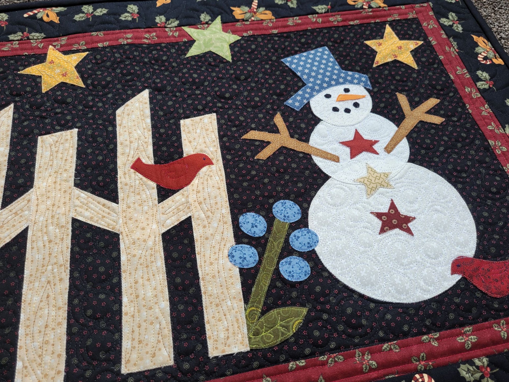 This applique snowman quilt is done in cotton fabrics with traditional prints of holly, candy canes, bows. The snowman has stick arms and the scene includes a rail fence, red birds, stars and flower on a black background with red border.