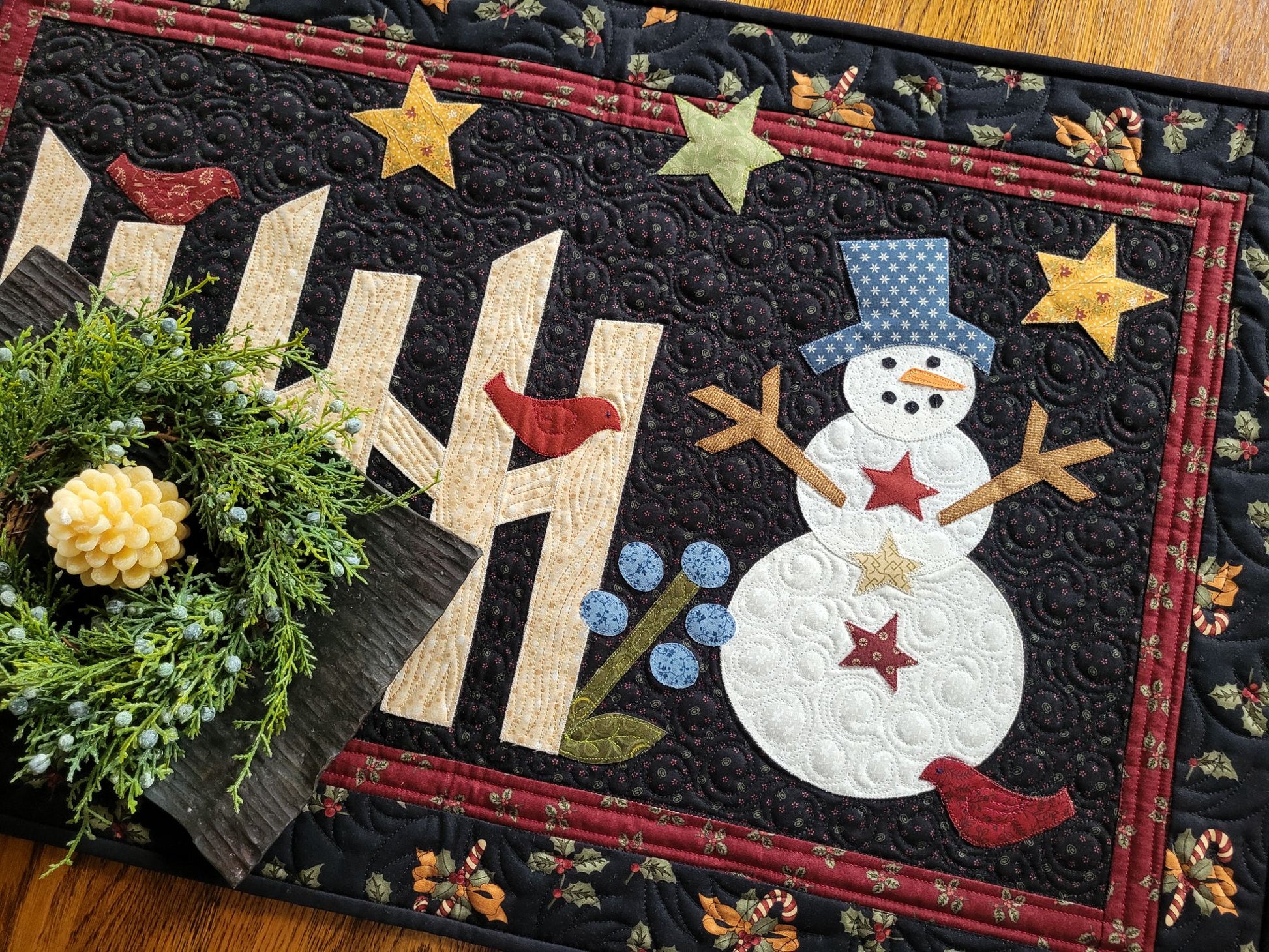 This applique snowman quilt is done in cotton fabrics with traditional prints of holly, candy canes, bows. The snowman has stick arms and scene includes rail fence, red birds, stars, flower on a black background with red border.