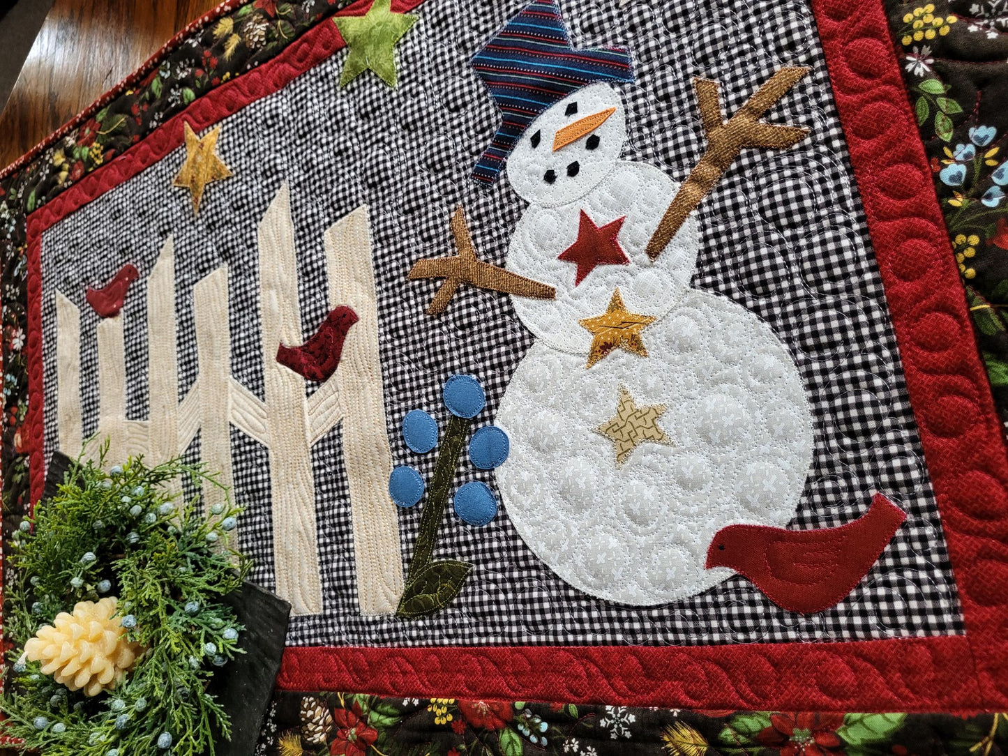 Cute snowman scene with rail fence, stars and birds on gingham background. The quilt has a small red border surrounded by black winter theme floral & pine cone print border.