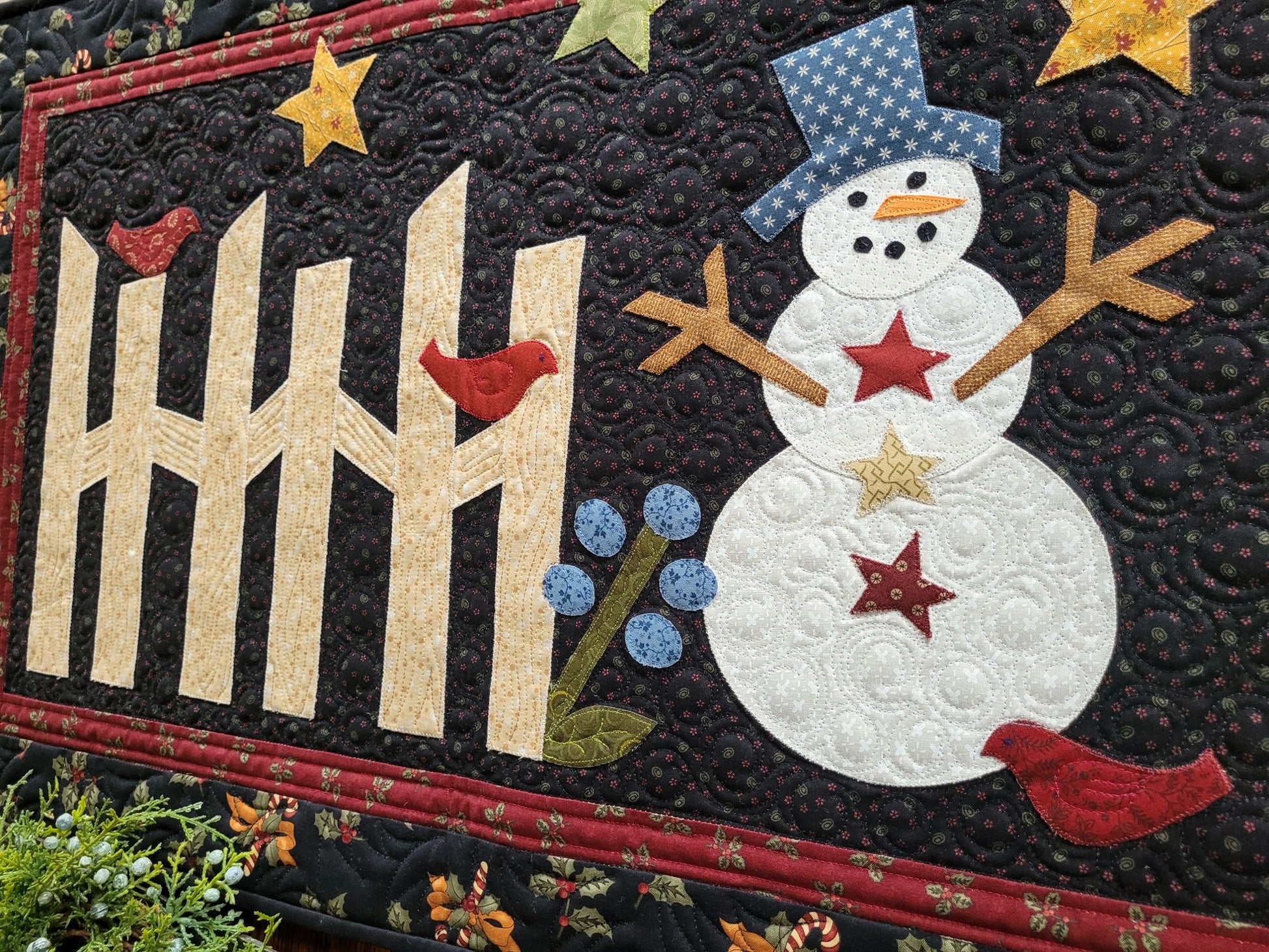 This applique snowman quilt is done in cotton fabrics with traditional prints of holly, candy canes, bows. The snowman has stick arms and scene includes rail fence, red birds, stars, flower on a black background with red border.