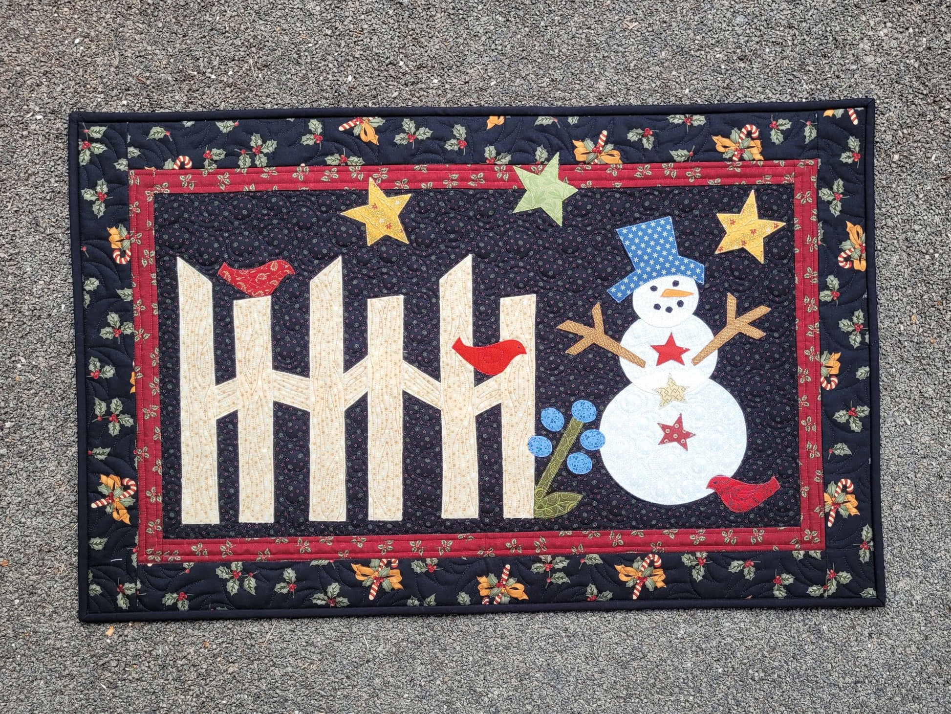 This applique snowman quilt is done in cotton fabrics with traditional prints of holly, candy canes, bows. The snowman has stick arms and the scene includes a rail fence, red birds, stars and flower on a black background with red border.