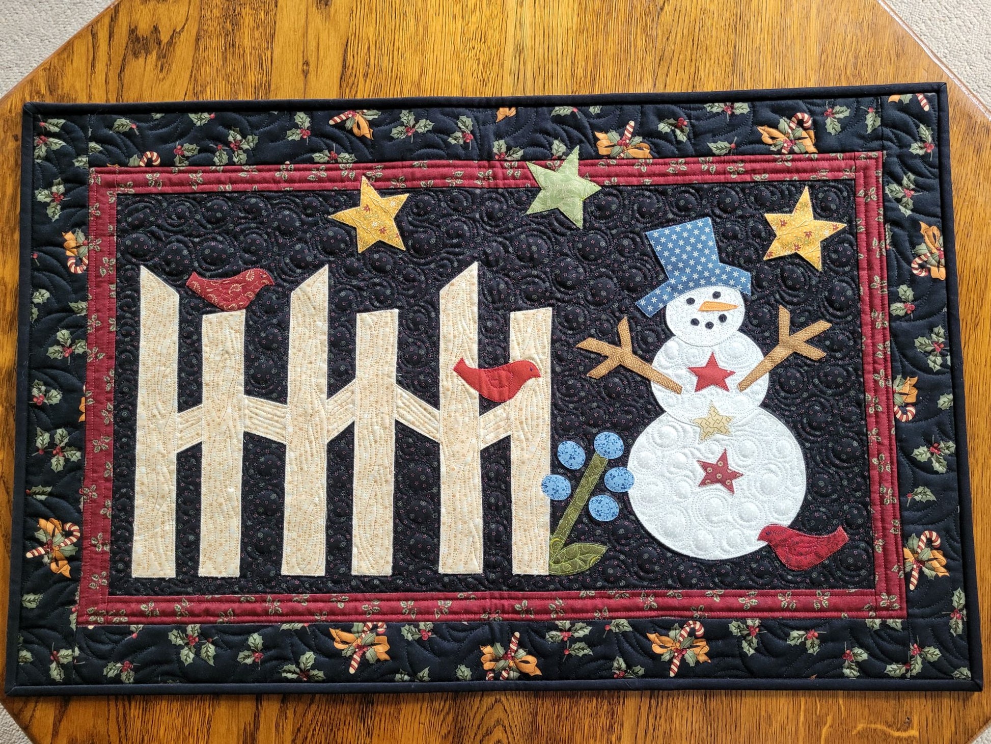 This applique snowman quilt is done in cotton fabrics with traditional prints of holly, candy canes, bows. The snowman has stick arms and the scene includes a rail fence, red birds, stars and flower on a black background with red border.