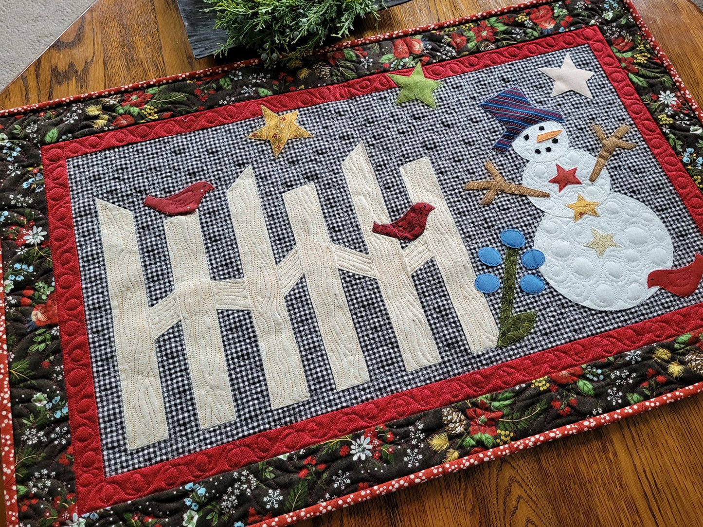 Winter snowman scene with rail fence, birds and stars are made with handcut applique shapes. Set on black and white gingham background and surrounded with red and black winter theme floral borders.