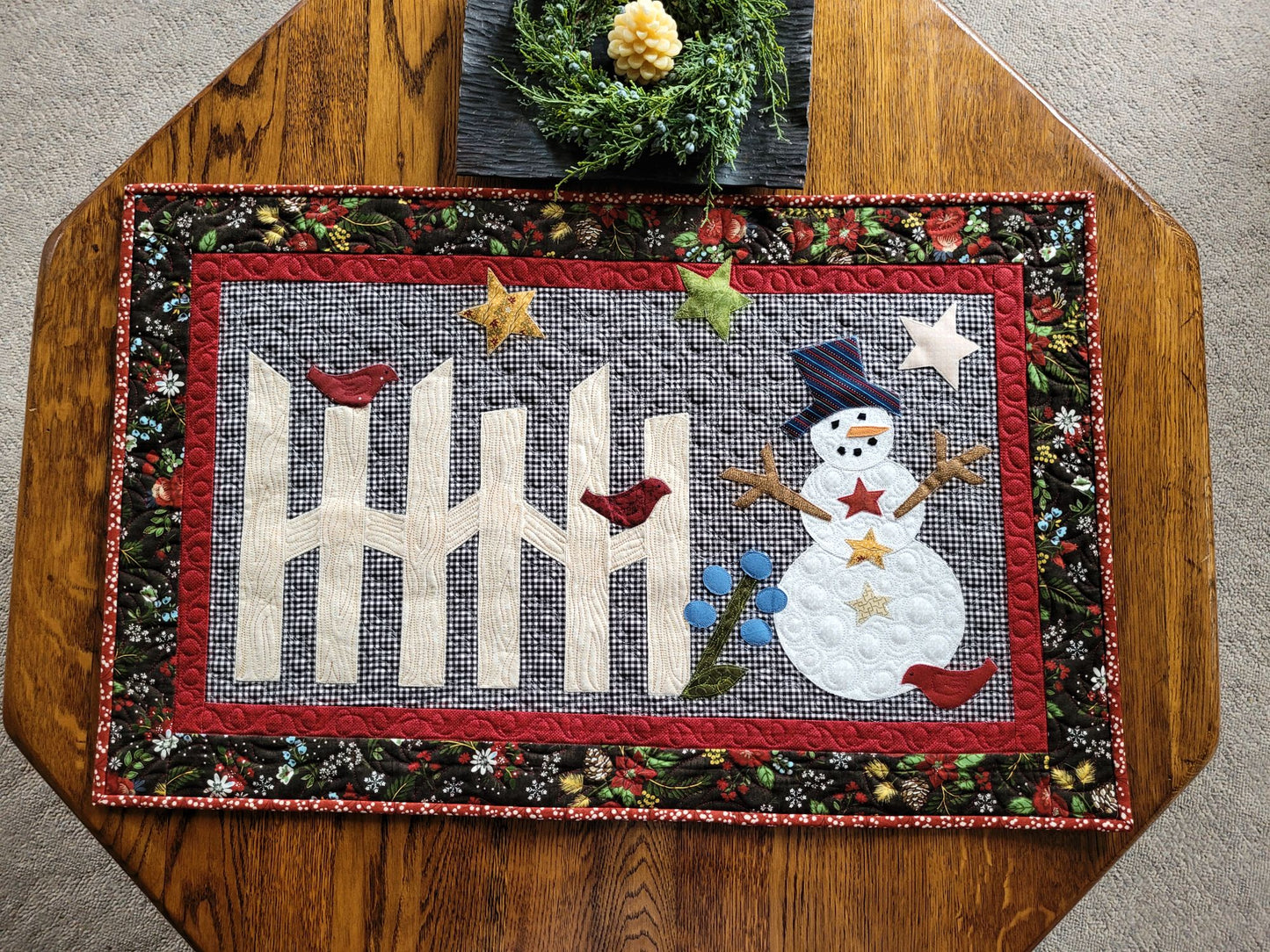 Cute snowman scene with rail fence, stars and birds on gingham background. The quilt has a small red border surrounded by black winter theme floral & pine cone print border. Quilts shown on hexagon wood table with candle.
