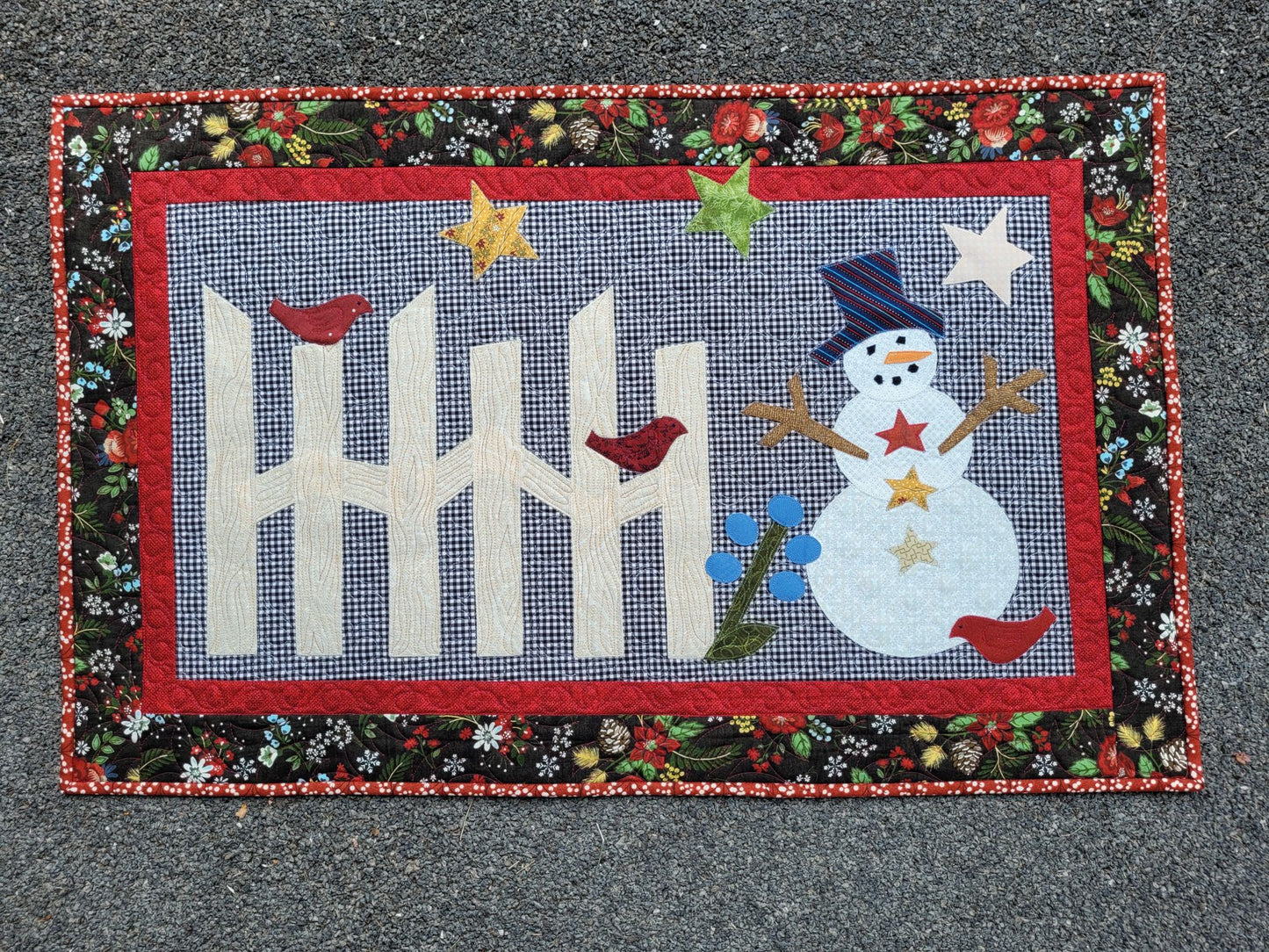 Cute snowman scene with rail fence, stars and birds on gingham background. The quilt has a small red border surrounded by black winter theme floral & pine cone print border.
