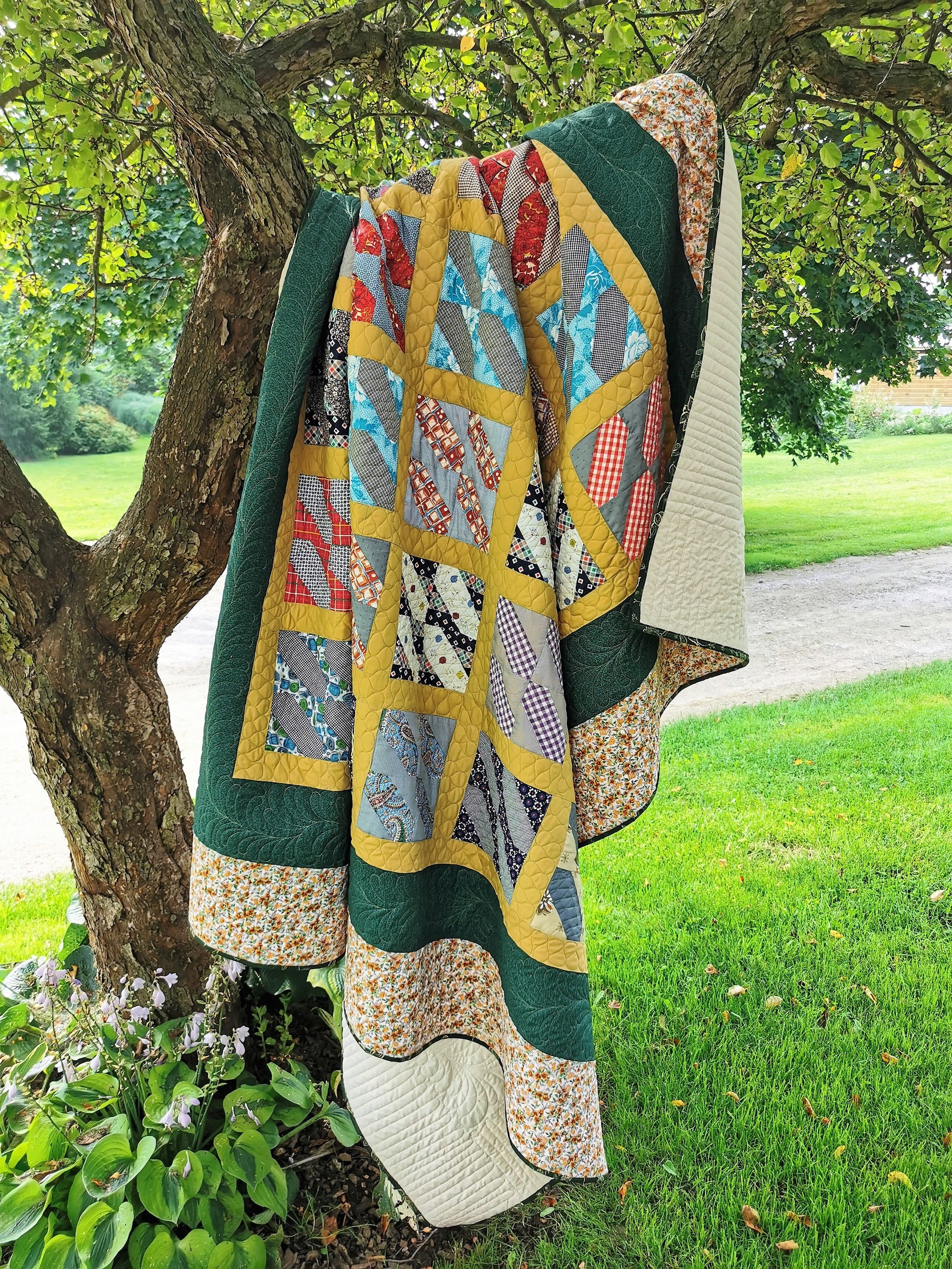 This large queen vintage bed quilt it shown in outdoor light, draped over the branch of a crab apple tree.  The middle of the quilt top has the vintage patchwork blocks set in a mustard yellow grid pattern. The interior grid is surrounded by a dark green border then a beige floral border around the outside