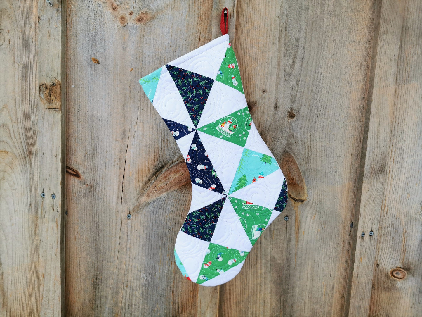 This heirloom quilted stocking has pinwheel piecing in winter themed fabrics on a white background. Pinwheels are navy, green and a light teal blue.