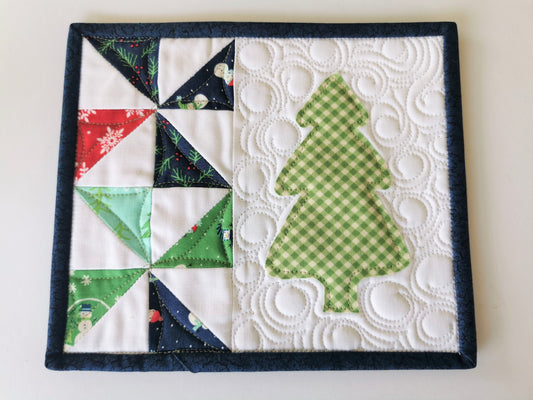 This mini christmas tree quilt features winter theme fabrics on white background. Scrappy pinwheel patchwork down the left side with a green gingham christmas tree on the right. Binding is a dark navy.