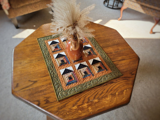 Decorative table quilt with quilted log cabin house design on a wooden table.