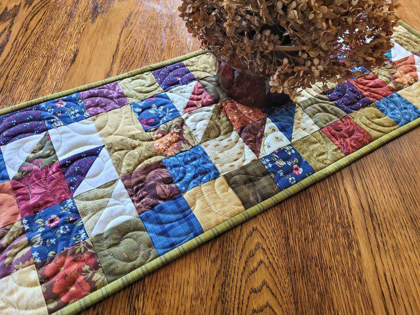 Quilted patchwork table runner on a wooden surface with a decorative item on top.