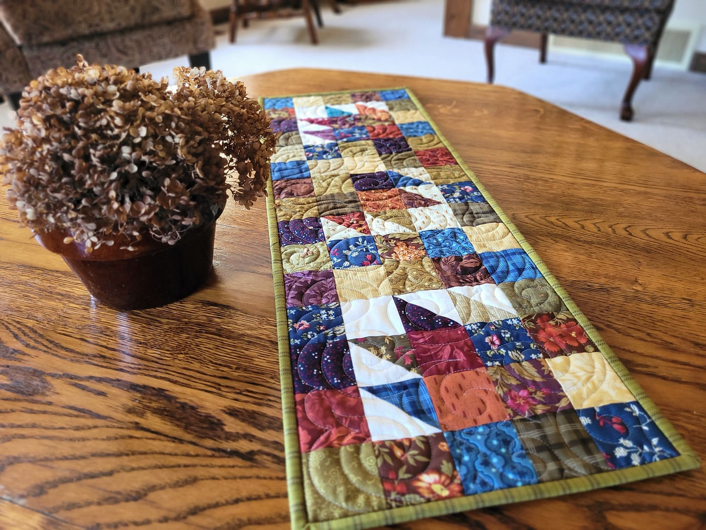 Colourful quilted table runner on a wooden table with a pot of dried flowers.