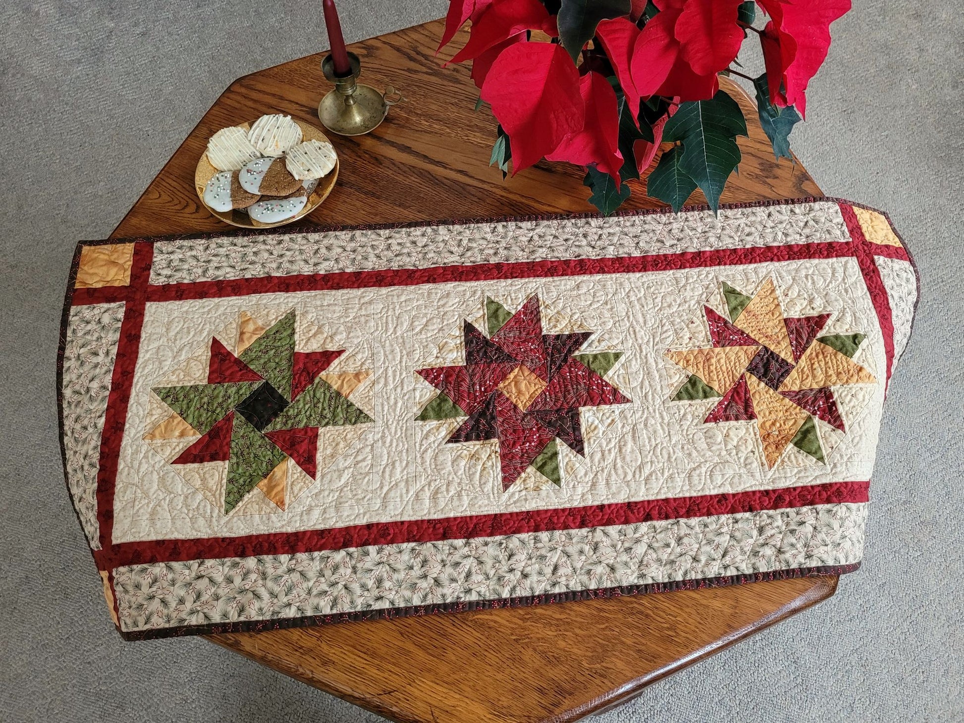 Quilted table runner with star patterns on a wooden table with a poinsettia, a plate of cookies and a candle.