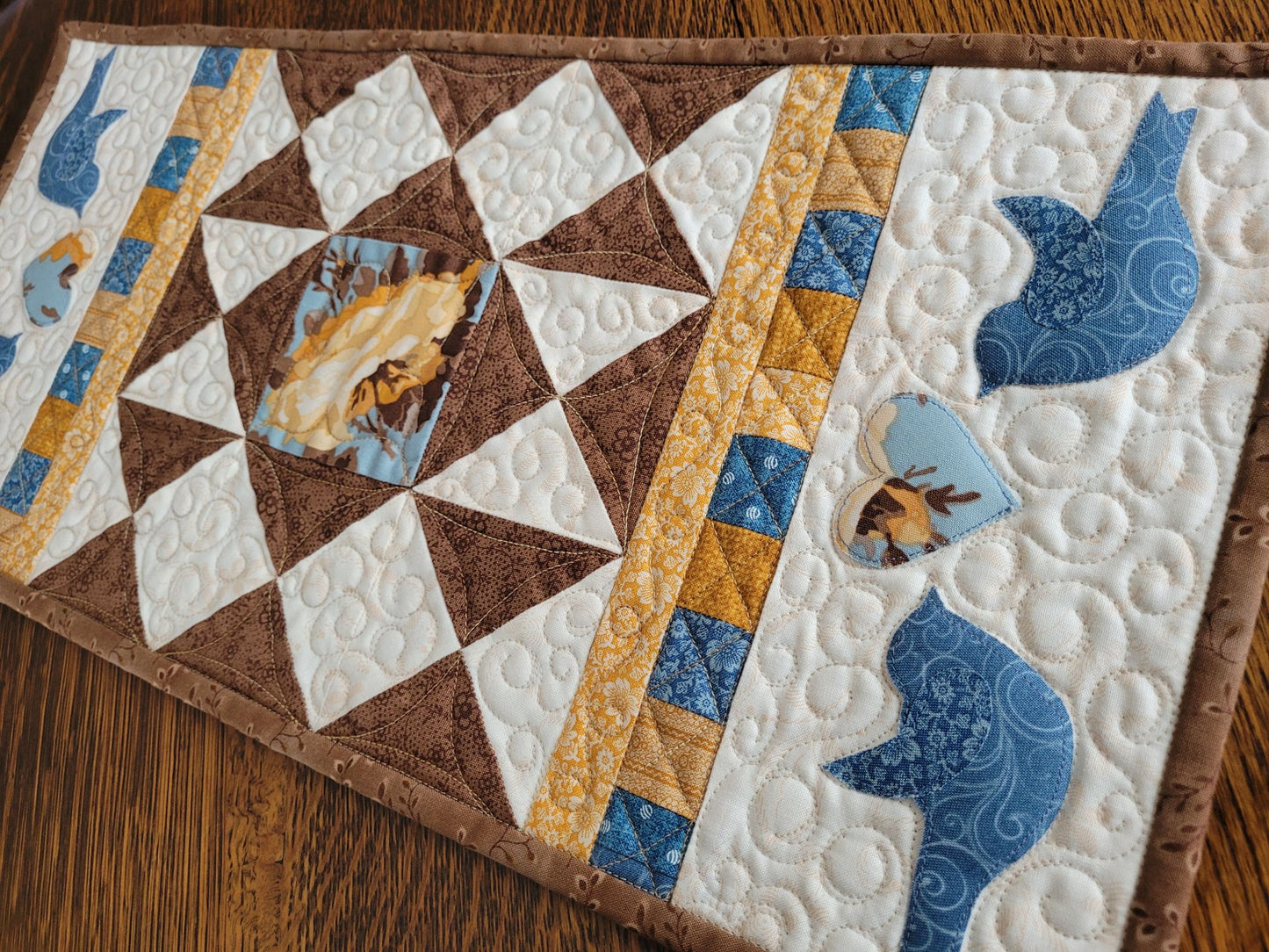 Small rectangle quilt with applique blue birds facing a floral heart at each end with brown patchwork in the centre. 