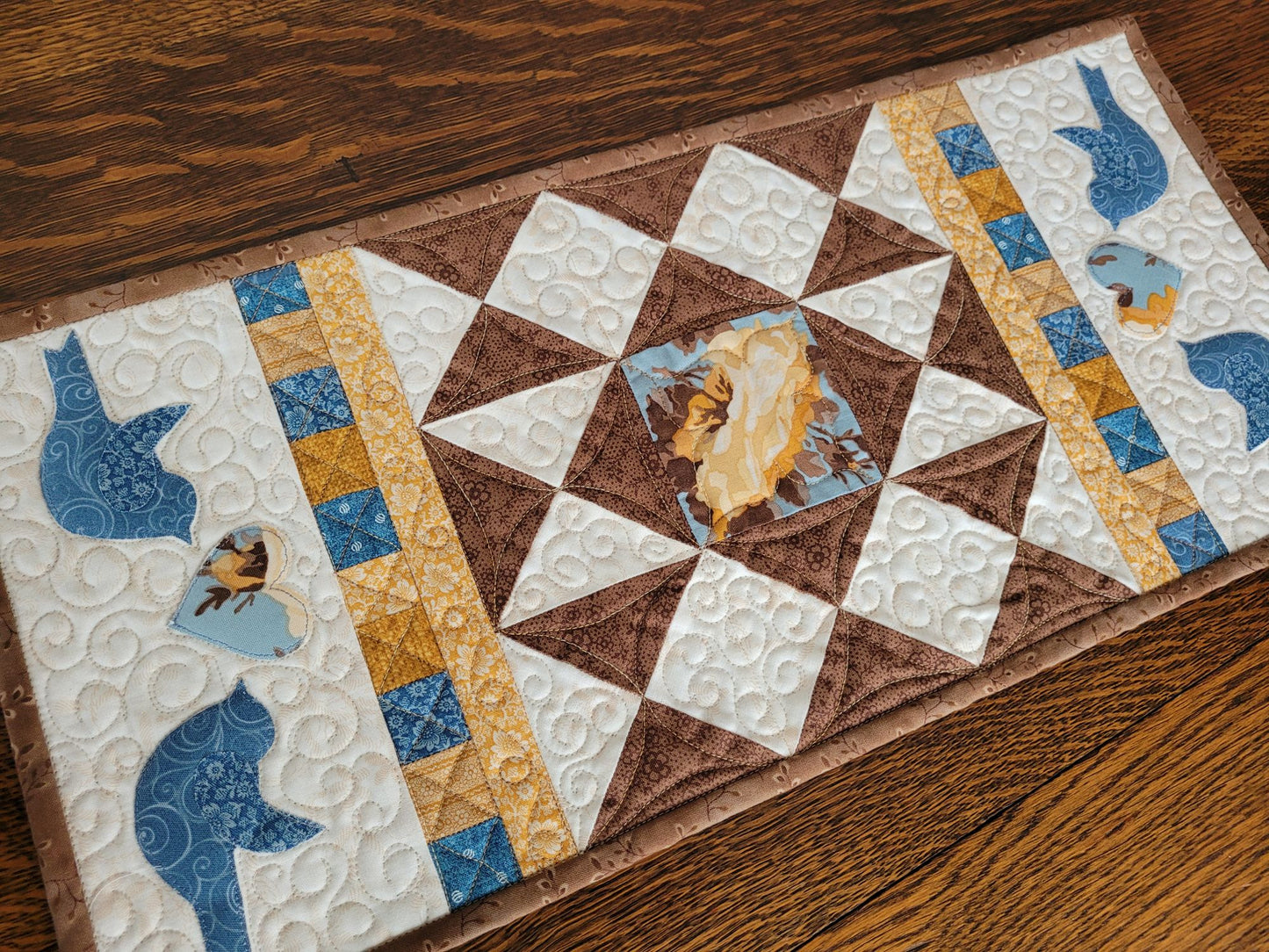 Quilted table runner in brown, blue and gold on a warm white background. Featured on each end is a set of blue birds flanking a floral heart.