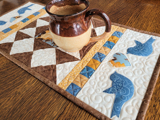 Quilted table runner with two appliqué blue birds on either side of a floral heart, on each end of the runner. Coordinating fabrics in brown, gold and blue on warm white background. Improv patchwork. 