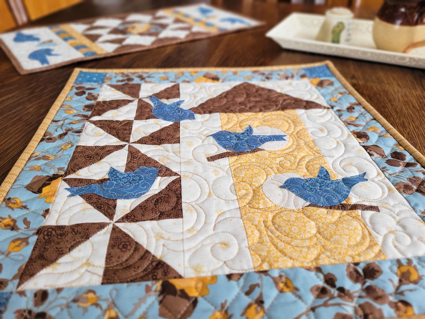 Thoughts of Spring Wall Quilt shown on a wooden table with the coordinating Love Birds Table Runner in the background.