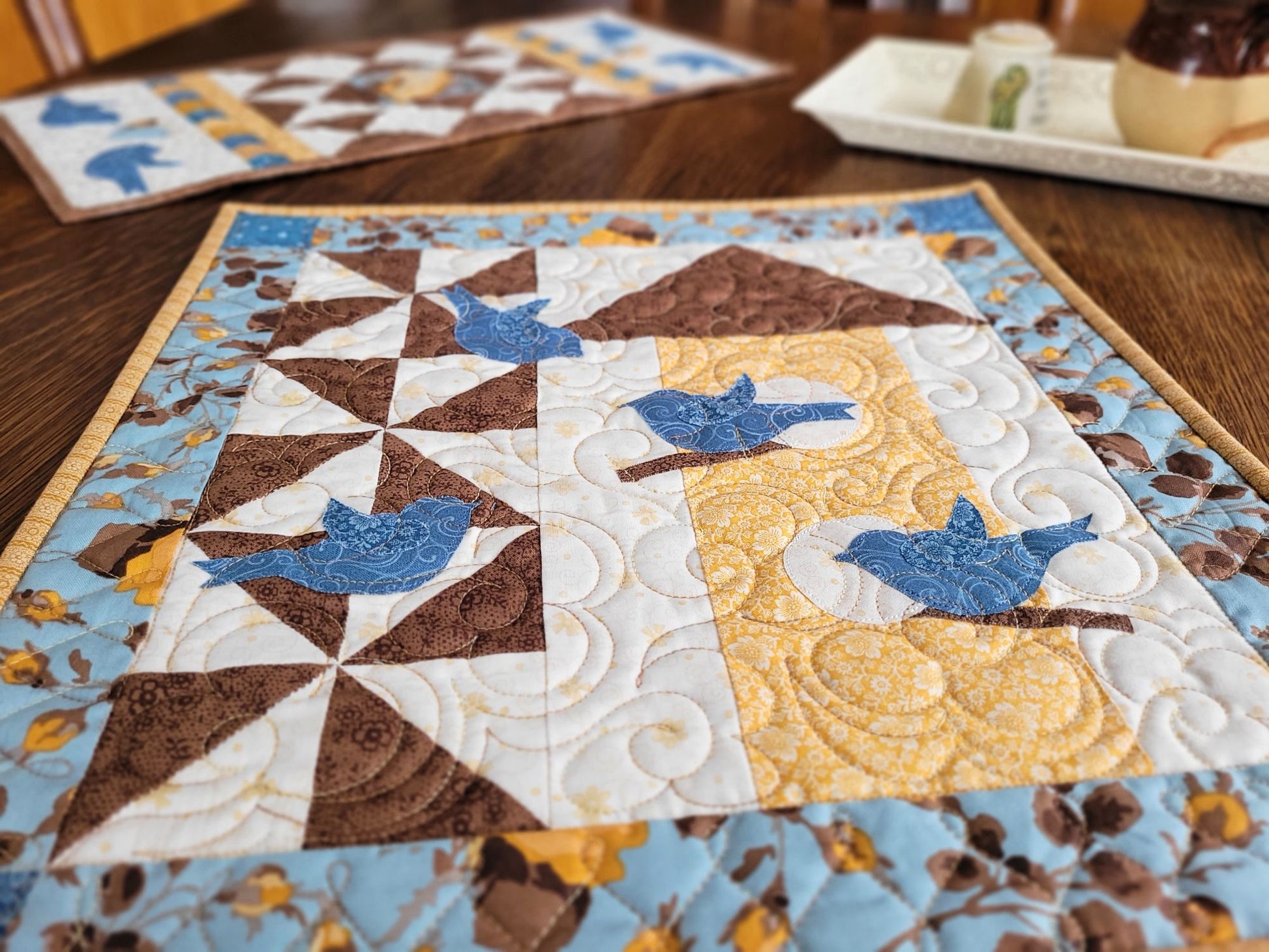 Thoughts of Spring Wall Quilt shown on a wooden table with the coordinating Love Birds Table Runner in the background.