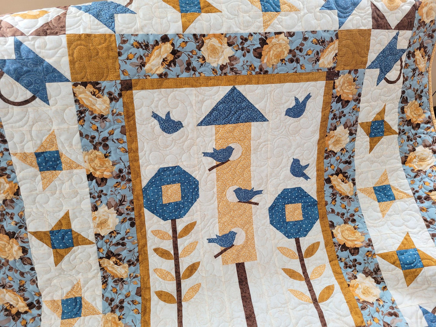 Showing close up of bluebird quilt with the birdhouse, patchwork flowers and stars surrounded by borders of roses on a sky blue background. 
