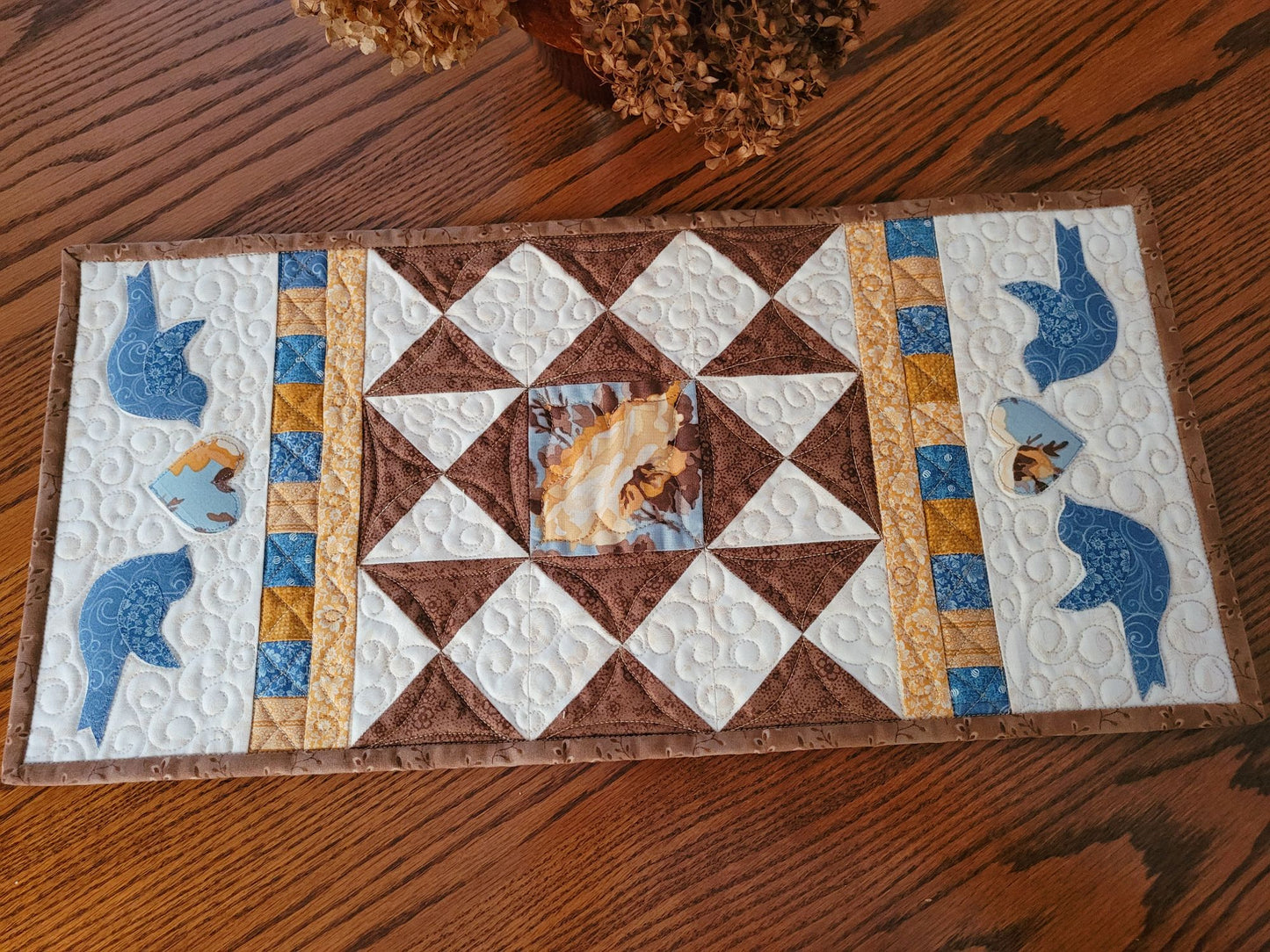 Quilted patchwork table runner on a wood table with dried flowers. On each end the runner has two sets of blue birds, each flanking a floral heart.  The warm white background shows off the other coordinating fabric colours of brown, blue, gold and yellow.