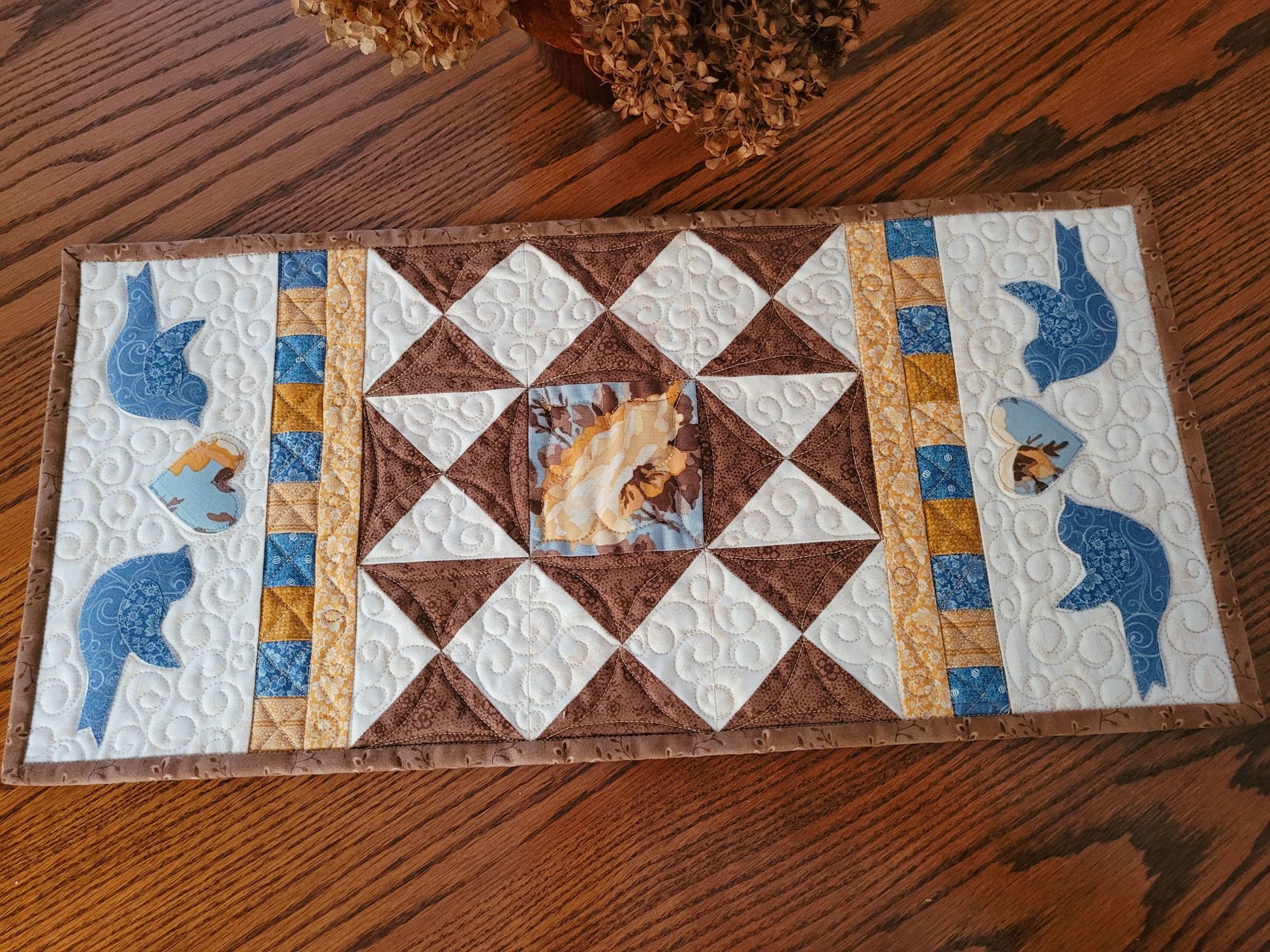 Quilted patchwork table runner on a wood table with dried flowers. On each end the runner has two sets of blue birds, each flanking a floral heart.  The warm white background shows off the other coordinating fabric colours of brown, blue, gold and yellow.