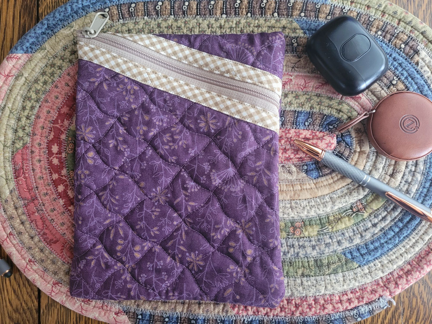 Purple quilted pouch with a zipper on a colourful woven mat with a pen, ear bud case and round tape measure case for scale.